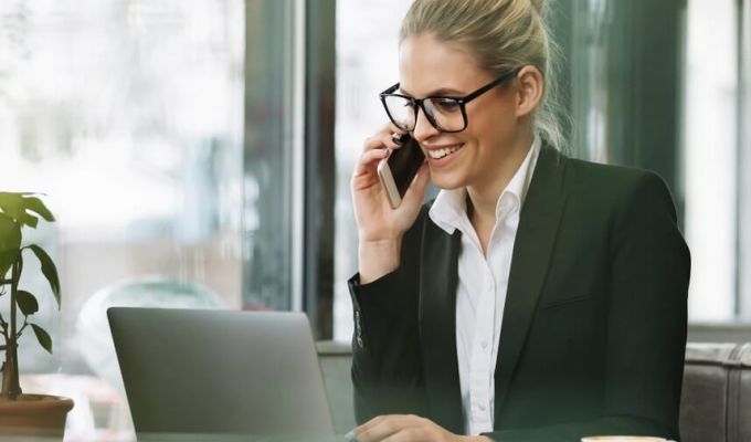 Kobieta o jasnych włosach rozmawia przez telefon i obsługuje laptopa. Ubrana jest w czarną marynarkę i białą koszulę. W tle jasne wnętrze i okna.