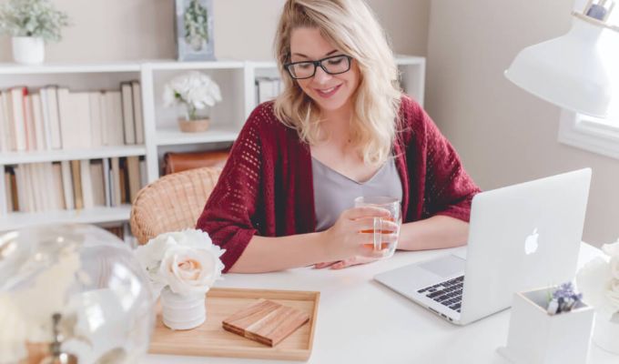 Kobieta siedząca przy biurku. Ma na sobie bordowy kardigan i szarą bluzkę. Kobieta ma blond włosy i okulary. Na biurku przed nią stoi laptop i szklanka z napojem. W tle widać białe półki z książkami