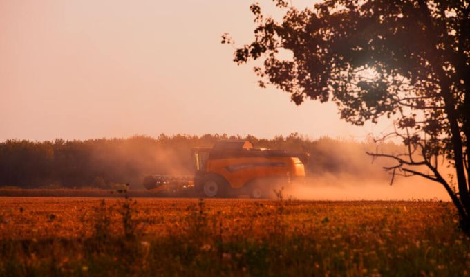 Kombajn pracujący na polu podczas zachodu słońca. W tle widać drzewa i las.