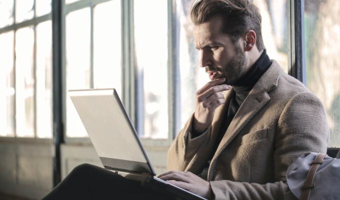 Mężczyzna ubrany w ciemny golf i beżowy płaszcz, siedzi, trzymając na kolanach otwarty laptop i zamyślony dotyka brody. W tle widać okno z białymi ramami, przez które wpada naturalne światło