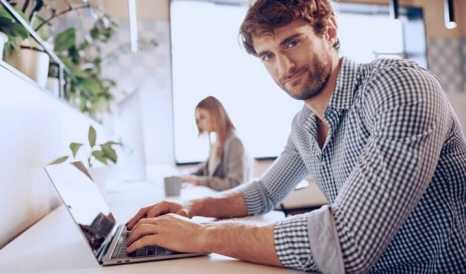 Mężczyzna w koszuli w kratę, pracuje na laptopie. W tle, widać kobietę, również pracującą przy komputerze. Znajdują się w jasnym, nowoczesnym biurze, z widocznymi roślinami doniczkowymi.