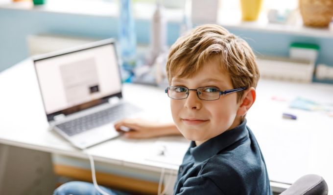 Uśmiechnięty chłopiec z krótkimi, brązowymi włosami i w okularach, który siedzi przy biurku. Na biurku przed nim stoi otwarty laptop.