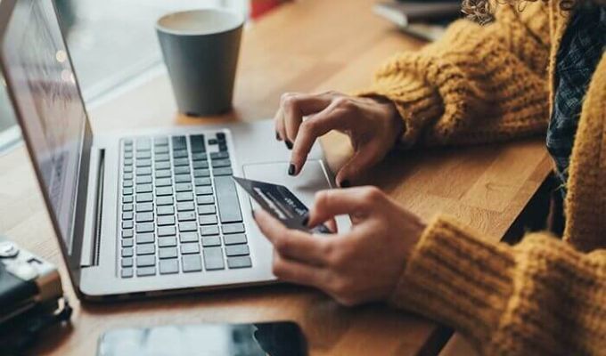 Osoba robiąca zakupy online. Kobieta trzyma kartę debetową w jednej ręce i używa laptopa drugą ręką. Na stole obok komputera znajduje się kubek i telefon. Kobieta ma na sobie żółty sweter.
