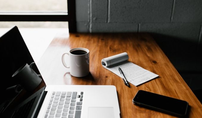 Drewniane biurko, na którym znajdują się: otwarty laptop, biały kubek z kawą, notes z długopisem i telefon komórkowy