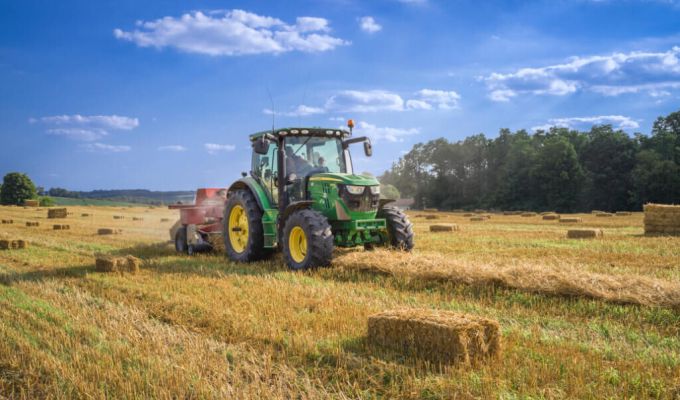 Zielony traktor, który pracuje na polu, zbierając siano. W tle widać kilka bel siana, a także las.